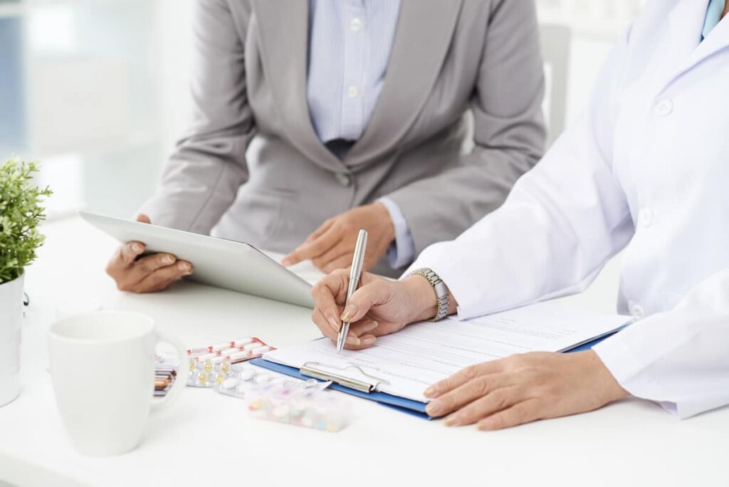A quality professional reviewing records on a clipboard beside packets of tablets while an auditor takes notes on a tablet.